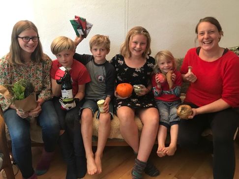Fünf Kinder und ihre Mutter sitzen auf einem Sofa und halten Schokolade, einen Kürbis, eine Mango und andere Lebensmittel aus der Biokiste in die Kamera.