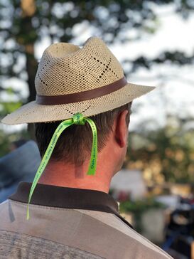 Ein Mann mit einem Hut von hinten. Am Hut ist ein hellgrünes Band mit einem Spruch befestigt.