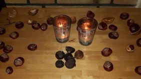 Zwei Kerzen stehen auf einem Tisch, ringsum verteilt liegen Kastanien. Direkt vor den Kerzen liegen sechs schwarze, schrumpelige Kastanien. 