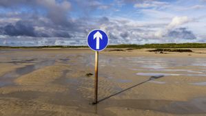 Ein Schild mit einem Pfeil, der geradeaus zeigt, im Sand. Hier geht es zu einer schottischen Insel, die nur bei Ebbe zu erreichen ist.