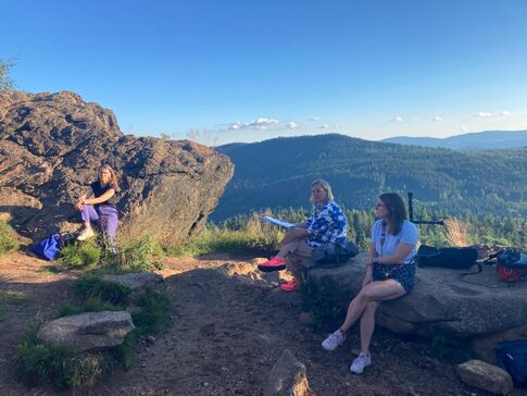 Menschen auf Baumstämmen auf dem Silberberg, dahinter Blick über die Berge und ins Tal.