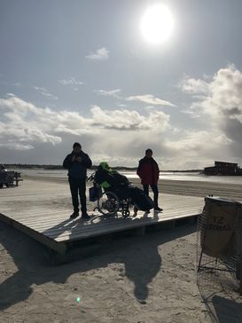 Eine Frau im Rollstuhl und zwei Begleiter stehen auf einem Strand vor einem leicht bewölkten Himmel im Gegenlicht.